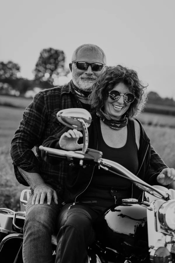 Man zit achterop de moter. Zijn vrouw zit voorop de motor. Beide zijn aan het lachen.